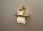 Gold toilet paper holder with a white roll and small potted plant on a brown background