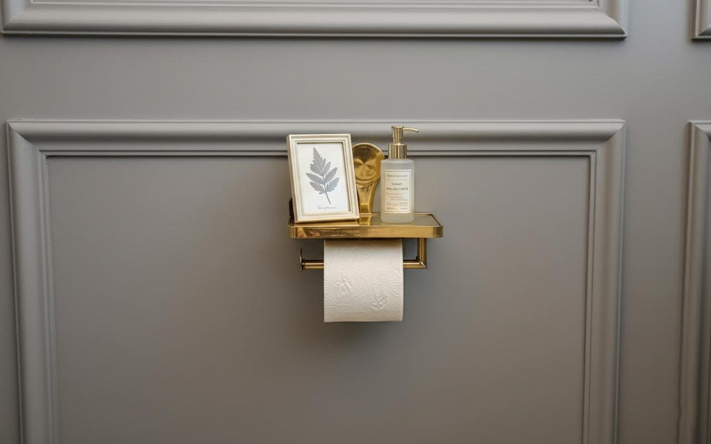 Gold toilet paper holder with a bottle and framed picture on a gray wall.