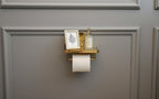 Gold toilet paper holder with a bottle and framed picture on a gray wall.