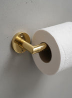 Gold toilet paper holder with a roll of toilet paper on a light gray wall.