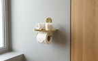 Gold wall-mounted shelf with toilet paper, candles, and a roll of paper towels against a light gray wall.