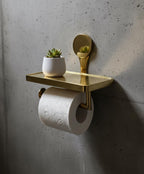 Gold toilet paper holder with a shelf, holding a roll of toilet paper and two small potted plants against a concrete wall.