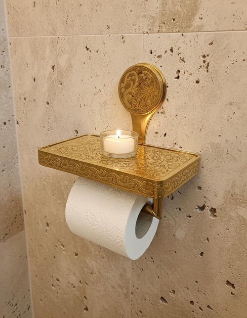 Decorative gold shelf with a candle and toilet paper holder on a beige tiled wall.